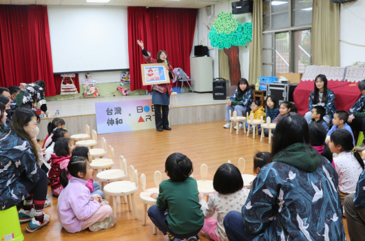 幼兒園教室內舉辦文化交流活動，小朋友圍坐成半圓形，講台上有人進行日語教學展示。牆上掛著台灣和日本的友誼旗幟，講台後方有投影幕和紅色布幕。教室布置溫馨，體現台日文化交流主題。