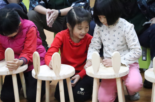 几名穿着红色和白色衣服的小女孩围坐在木制椅子周围，专注地参与室内活动。她们看起来在进行集体游戏或教育活动，身后有其他儿童和成人在场。场景温暖而充满童趣。