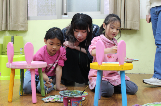 一位成人和兩名兒童在教室內進行美術活動。他們蹲在地上，共同組裝粉紅色和彩色的積木結構。地板上散放著美術用品和顏料盤。背景是綠色牆面和窗戶。
