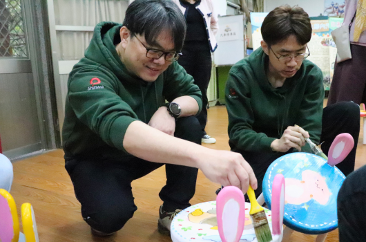 兩位穿著綠色運動衣的學生在室內活動中蹲下操作彩色手工藝品，旁邊擺放著黃色和粉紅色的裝飾物品。背景有其他參與者和教室環境。