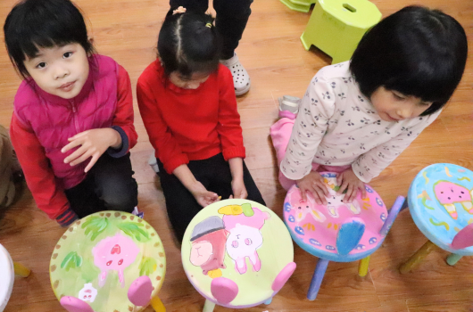 三名幼童蹲坐在木質地板上，各自手持彩色兒童椅。左起穿著紅色和黑色服裝，中間穿紅色衣服，右邊穿淺色衣服。椅子上印有卡通圖案，包括粉紅豬小妹等角色。背景有綠色物品和玩具。