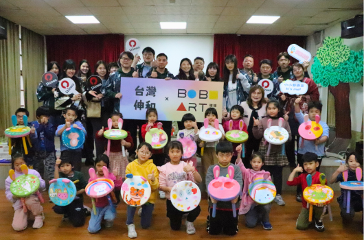 台灣伸筆與日日合作的兒童美術活動現場。約四十位穿著制服的小學生和教師聚集在室內，展示自己繪製的彩色紙盤作品。背景掛著活動主題布條，牆上有綠色樹木裝飾，呈現溫馨的教育活動氛圍。