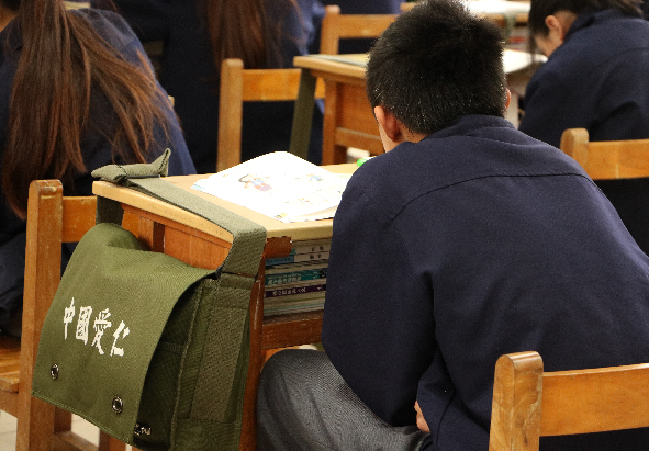 教室內，學生穿著制服坐在木製課桌椅上認真讀書。桌上放著開啟的課本和綠色書包，背景有其他同學也在學習。光線柔和，營造出寧靜的校園學習氛圍。