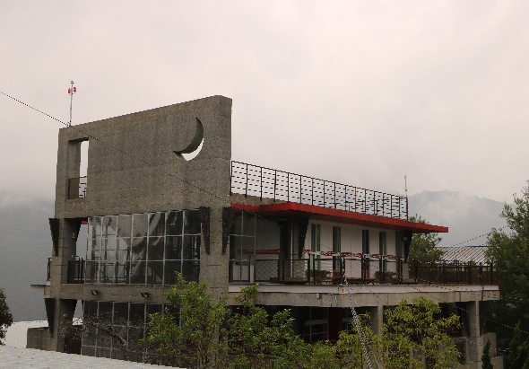 一棟現代建築物矗立在湖邊，混凝土外牆上裝有月牙形窗口，建築物分多層設計，配備玻璃窗和紅色橫樑，周圍環繞著綠色植被，背景為霧氣籠罩的山峰。