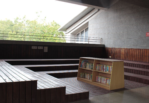 現代建築室外平台區域，木製階梯和地板面向綠樹背景。木製書架放置在紅磚地面上，展示各類書籍。灰色建築牆面配有玻璃窗和金屬欄杆，營造開放式閱讀空間。