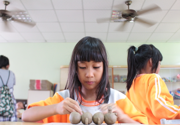 小學教室內，穿著橙色運動服的女童正專注地用手指塑造黏土球體，桌上擺放多個黏土作品。其他同學也在進行美勞課程，教室天花板裝有風扇和燈具。