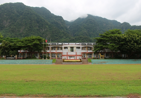 一所學校的校園全景，綠色足球場前方，白紅相間的校舍建築物配有藍色圍欄，背景是蒼翠的高山，山頂雲霧繚繞，校園周圍植有大樹，呈現寧靜的山區學校風景。