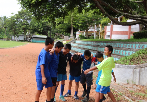 一群穿著藍色和黃綠色運動服的學生在紅土運動場上圍成一圈，進行團隊活動或訓練。背景是學校建築、濃密樹木和藍白相間的圍欄，陽光灑落在草坪上。