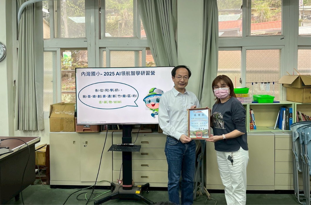 室內場景，兩位成人在教室內舉著獎狀合影。身後顯示屏展示「內灣國小 2025 AI 航智學研習營」海報，含卡通人物及中文文字。教室內可見書架、窗戶及教學用品。