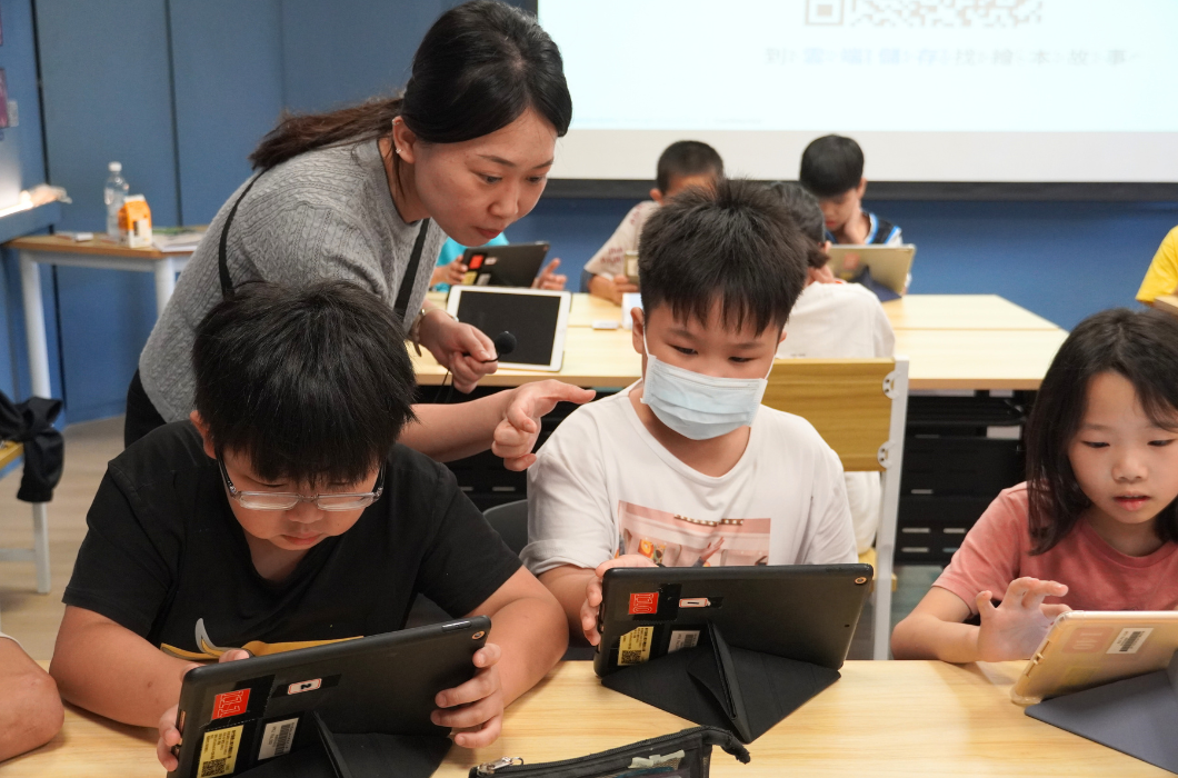 教室內，一位女性教師站在身後指導，幾名小學生坐在木製課桌前，各自操作平板電腦進行學習。一名學生戴著口罩，其他學生專注於螢幕。背景有投影幕和藍色牆面，呈現互動式數位教學的場景。