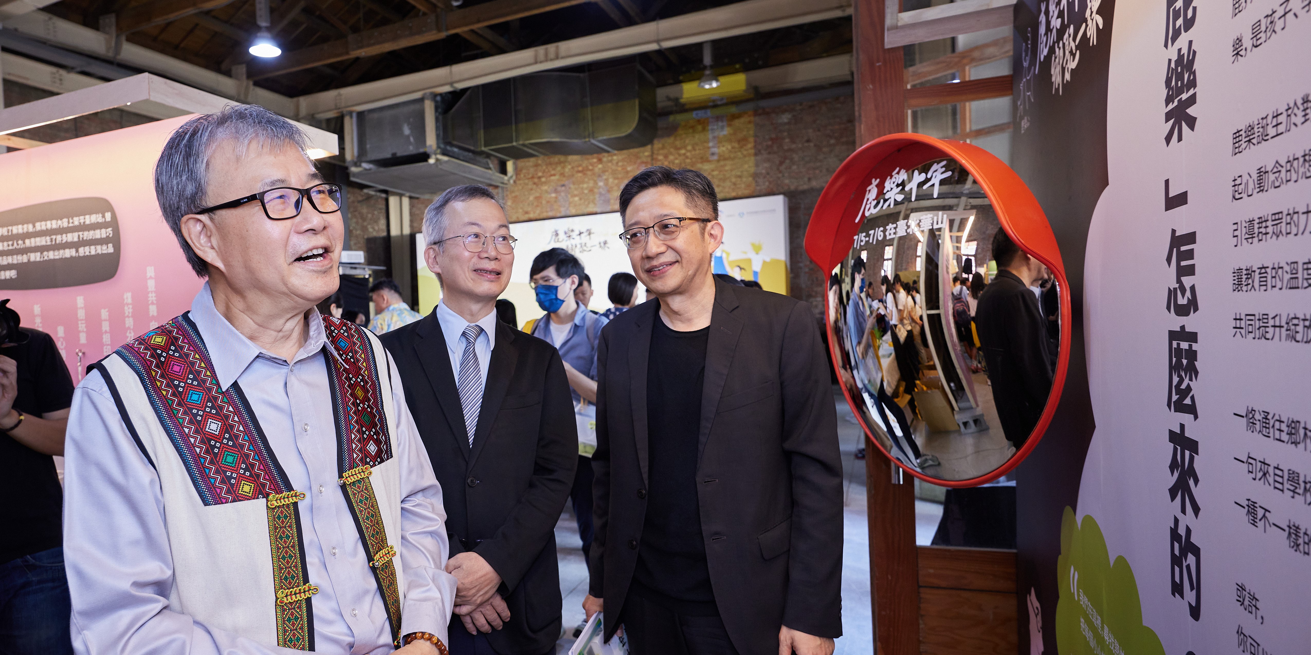 三位男性在室內展覽場景中交談，左方男子穿著傳統刺繡背心，中間和右方男子著正式西裝。右側紅色圓形框內展示活動照片，背景有黑色展板和中文文字，呈現正式的展覽或活動現場。