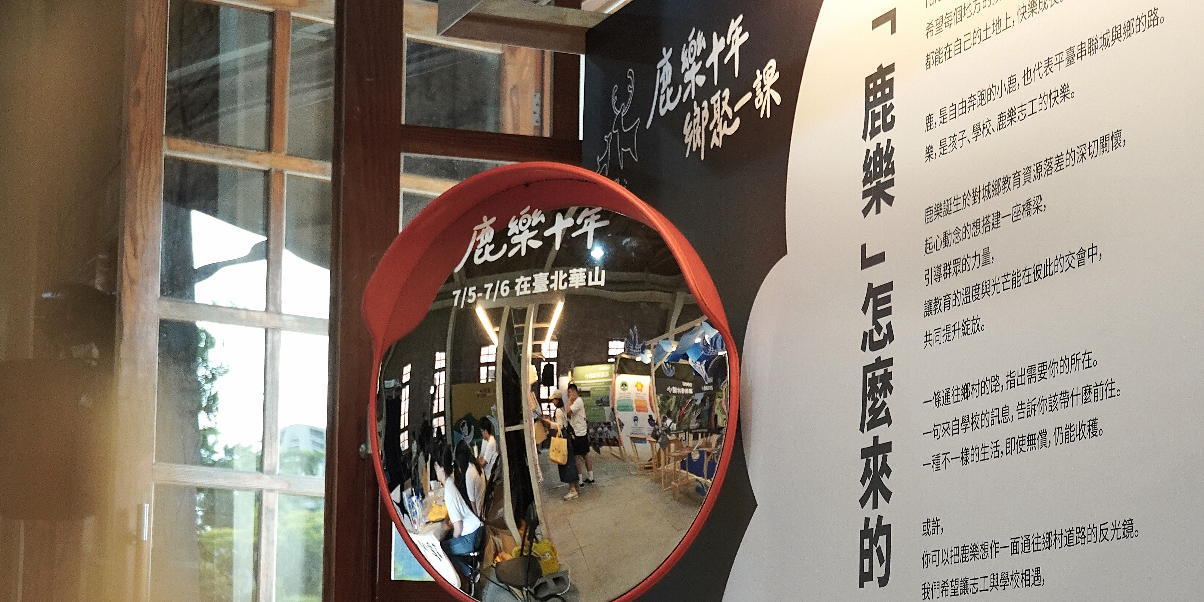 室內展示空間，牆上有黑色背景的白色中文文字和鹿樂標誌。紅色圓形鏡子映照出展覽現場，可見多位訪客參觀。右側白色牆面上有繁體中文段落文字。左側木製窗框透出室外景觀。