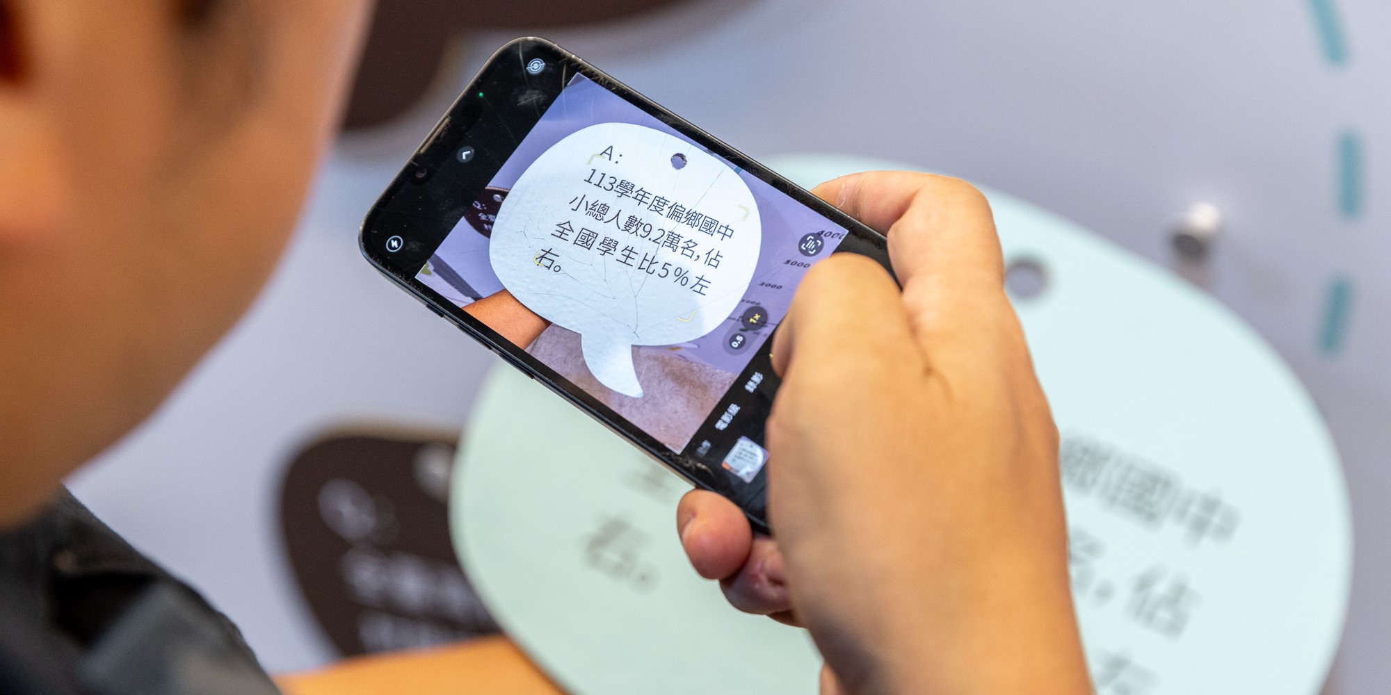 一個人手持黑色手機，拍攝圓形白色標籤上的繁體中文文字。標籤上顯示「113學年度偏鄉國中小總人數與2班至」等內容，下方為圓形圖表。背景為模糊的室內環境。