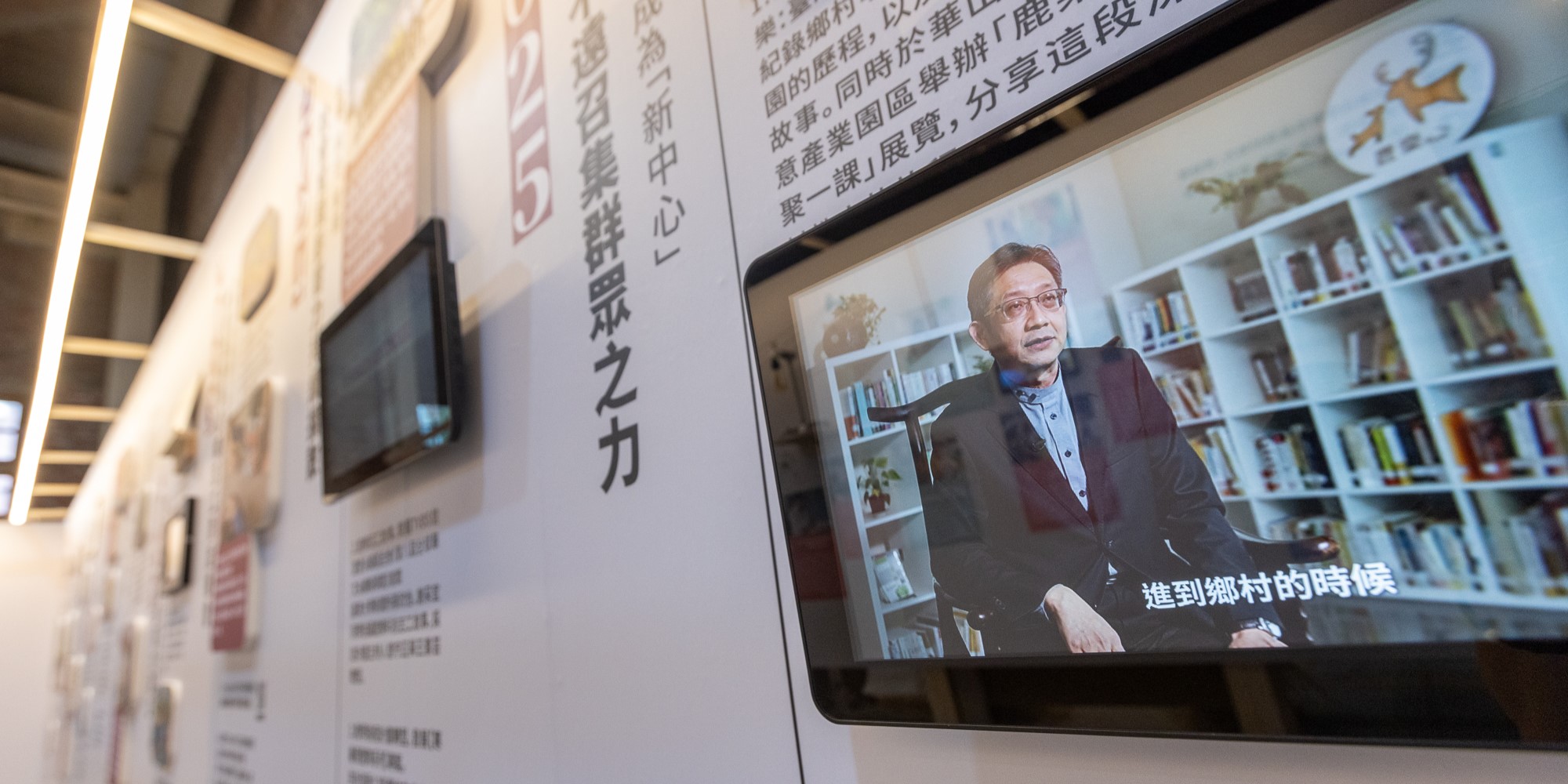 室內展示空間，牆面貼有繁體中文標語，右側大屏幕播放一位戴眼鏡的中年男性在書房內接受採訪的視頻，屏幕下方顯示中文字幕，左側牆面有多個小屏幕展示相關內容，天花板配備線性燈具。