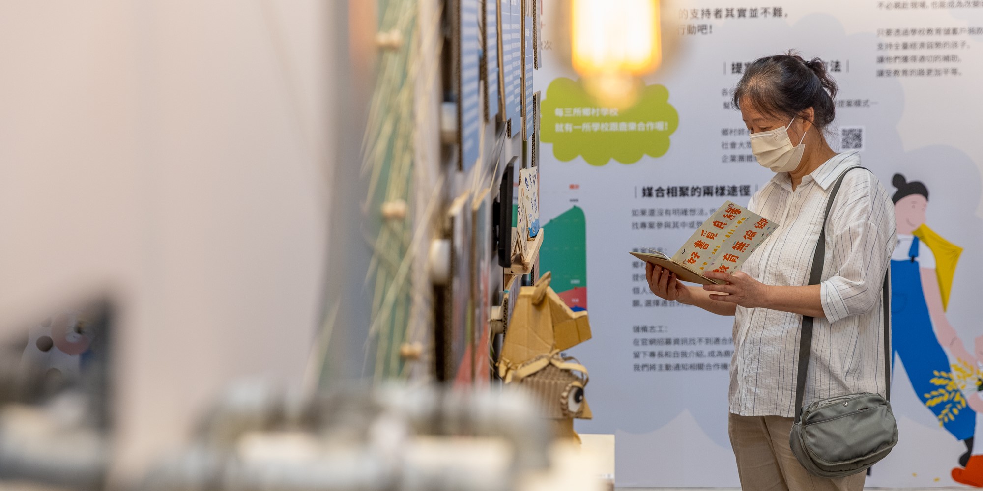 一位戴著口罩的女性在展覽會場閱讀手中的書籍。身後是色彩繽紛的展示牆，上面有綠色與黃色的設計元素、中文文字說明和圖示。牆邊擺放著黃色的裝飾品。場景顯示這是一場教育或文化主題的展覽活動。