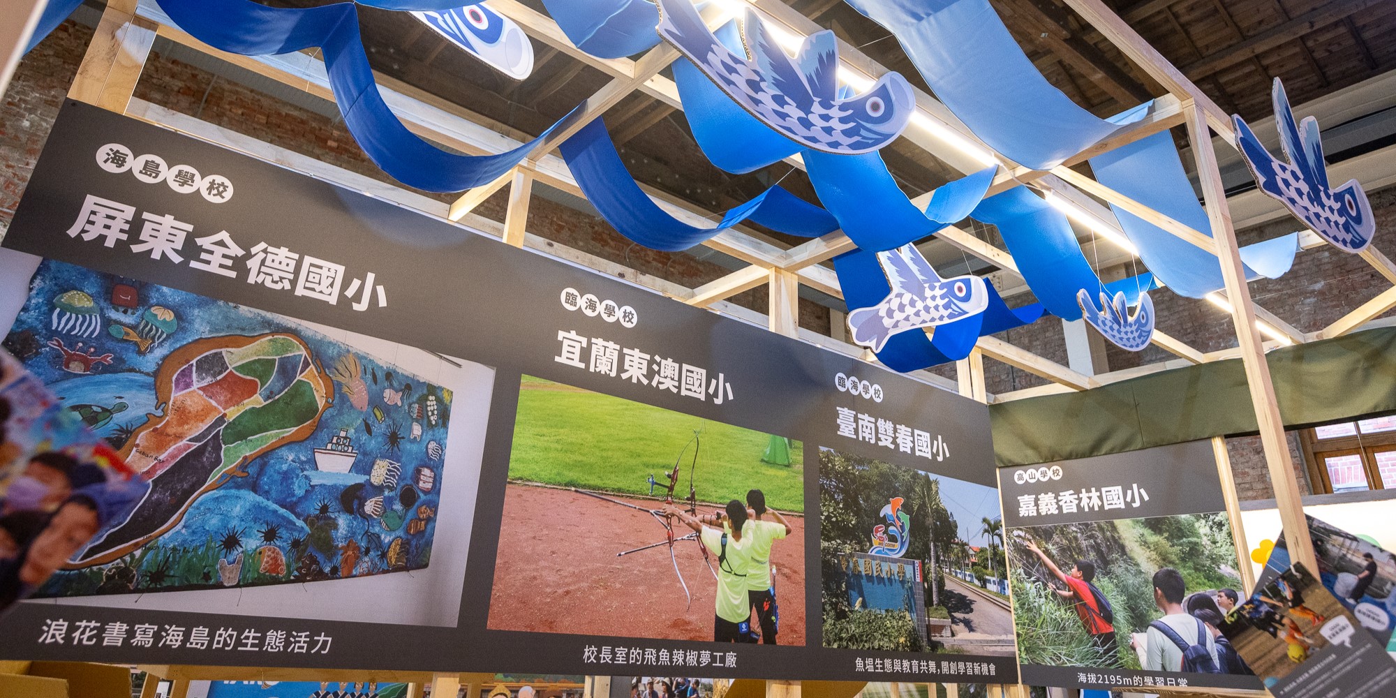 室內展覽展示三所學校的特色教育成果。黑色展板上方懸掛藍白色魚形燈飾，展板展示屏東全德國小的海島生態藝術、宜蘭南澳國小的射箭課程、台南雙春國小的森林教育等校園活動照片和學生作品。