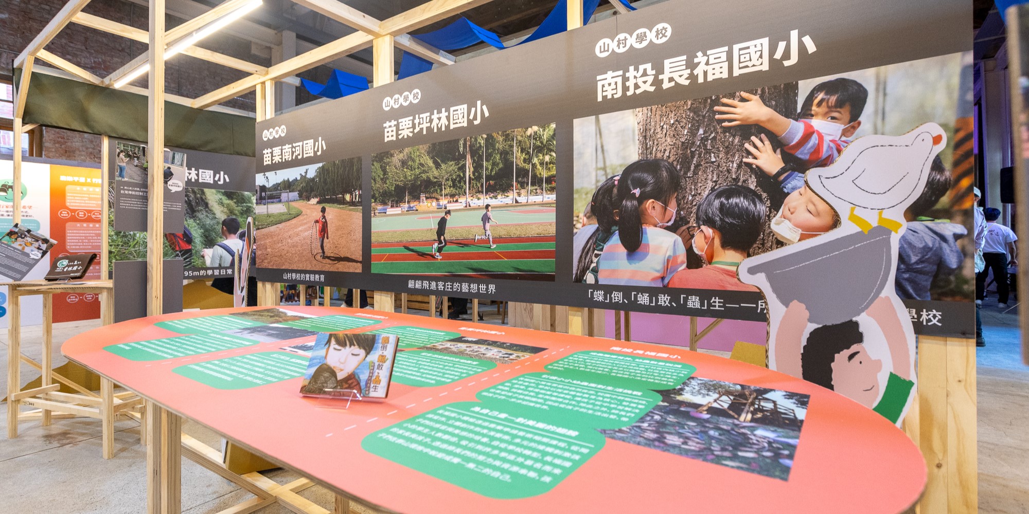 展示台灣小學的教育展覽展台。粉紅色互動展示桌展示苗栗坤林國小、苗栗南投國小和南投長福國小的校園照片及資訊。背景黑色面板呈現學生運動、樹木擁抱和戶外教學的場景。左側有橙色資訊看板。木製結構框架搭配彩色設計元素。