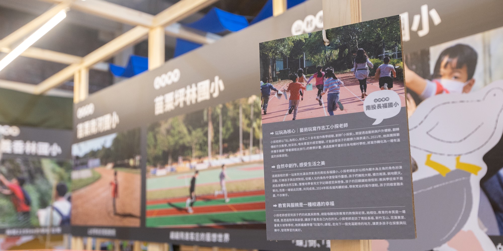 展示學校運動場景的宣傳板，左側顯示兒童在紅色跑道上奔跑的照片，右側為白色文字說明牌，介紹學校的教育理念與運動設施，背景為現代建築和藍色天花板。