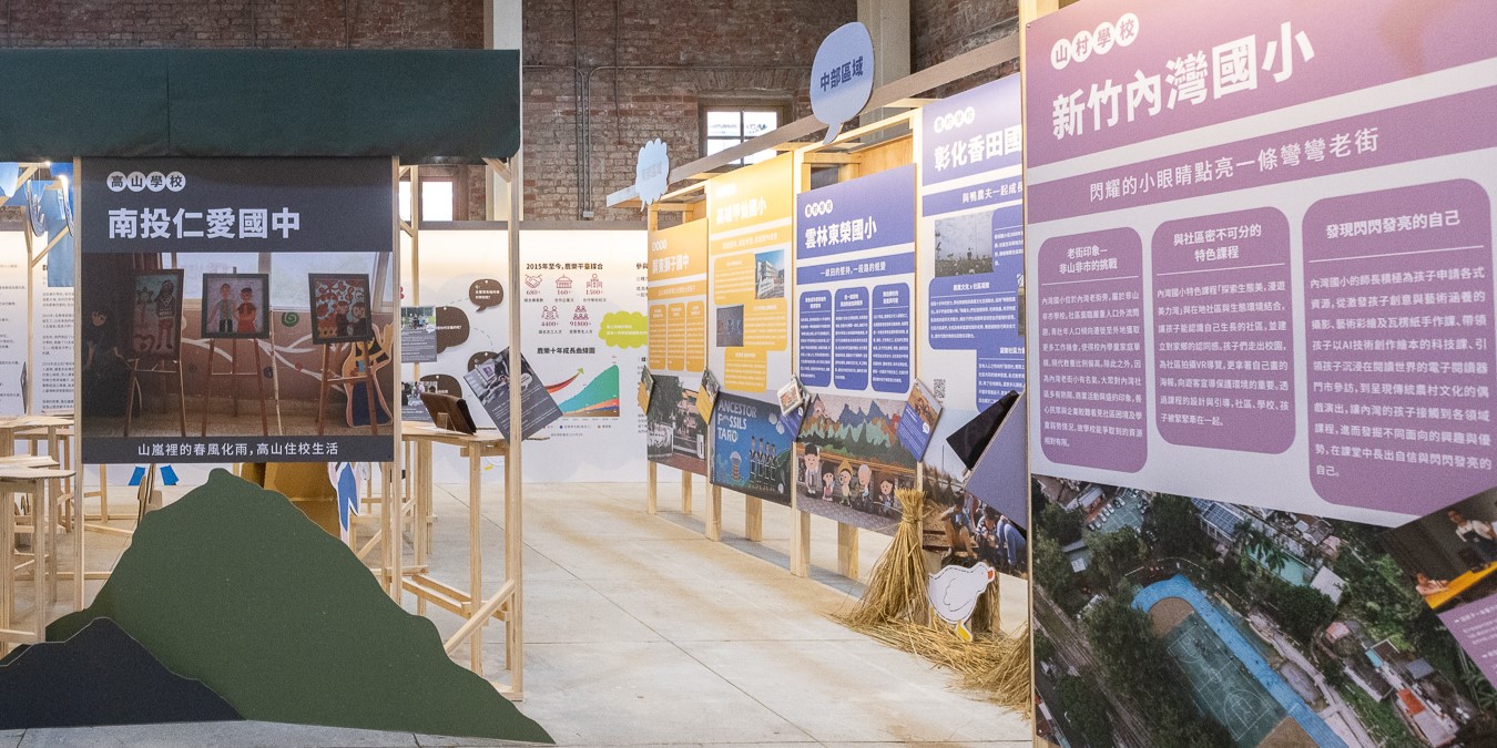室內展覽空間展示多個中文海報展板，介紹台灣地區的城鎮發展與社區特色。展板內容包含文字、照片和圖表，地面鋪設綠色裝飾造型，展現地方文化與城市規劃主題。