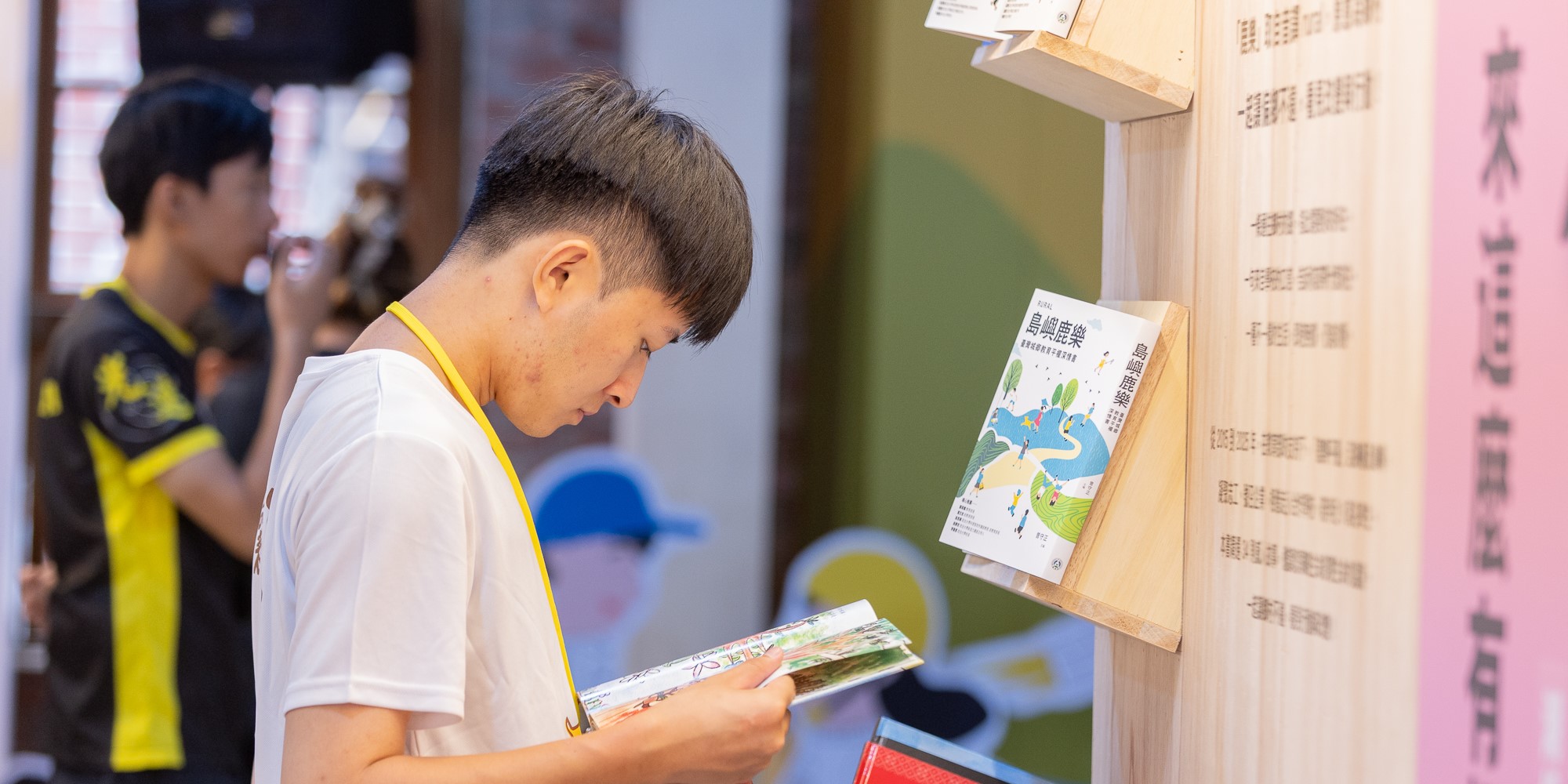 一名穿著白色上衣、掛著黃色識別證的男童在室內展覽中閱讀手中的宣傳冊，身旁木製展示板上展示著多份宣傳品，背景可見其他參觀者在展覽空間內活動。