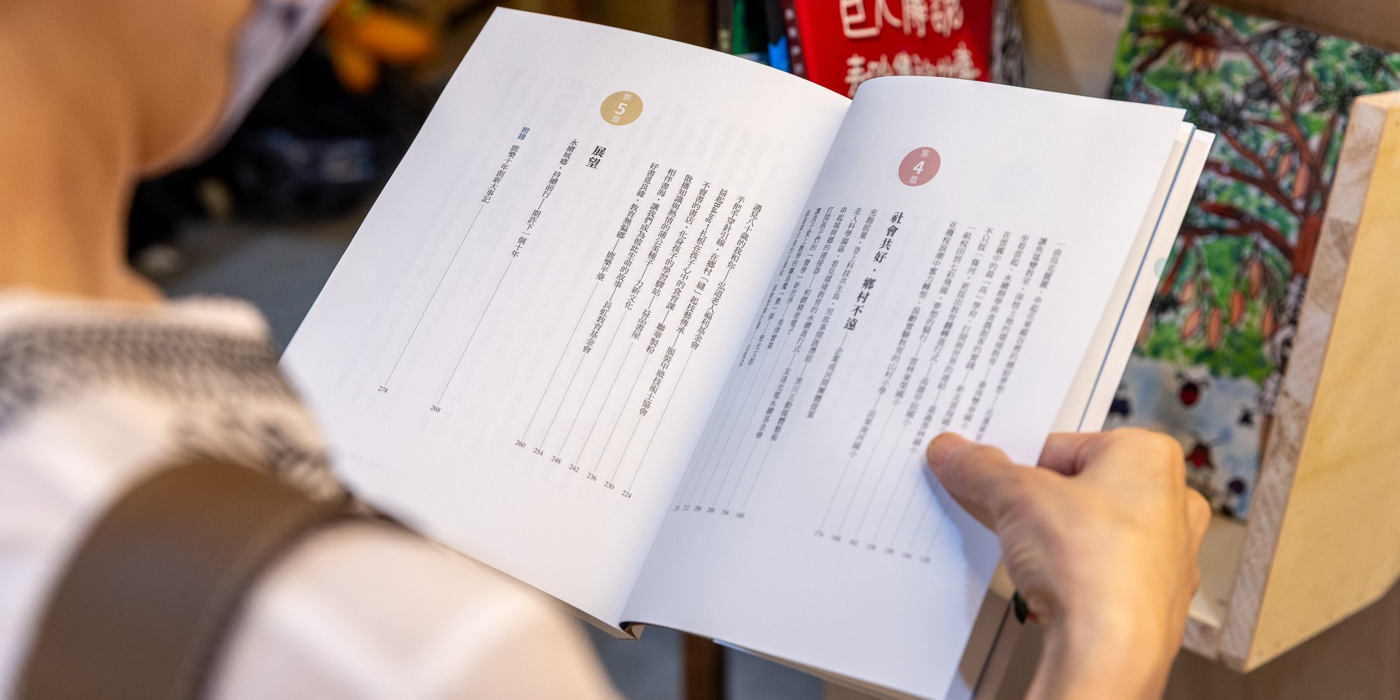 一個人正翻閱一本開啟的書籍，展示第五章的目錄頁面。左頁列出多個小節標題和頁碼，右頁則顯示章節內容。書籍採用簡潔的白色版面設計，頁面上有圓形的章節標號。背景可見其他物品和裝飾品。