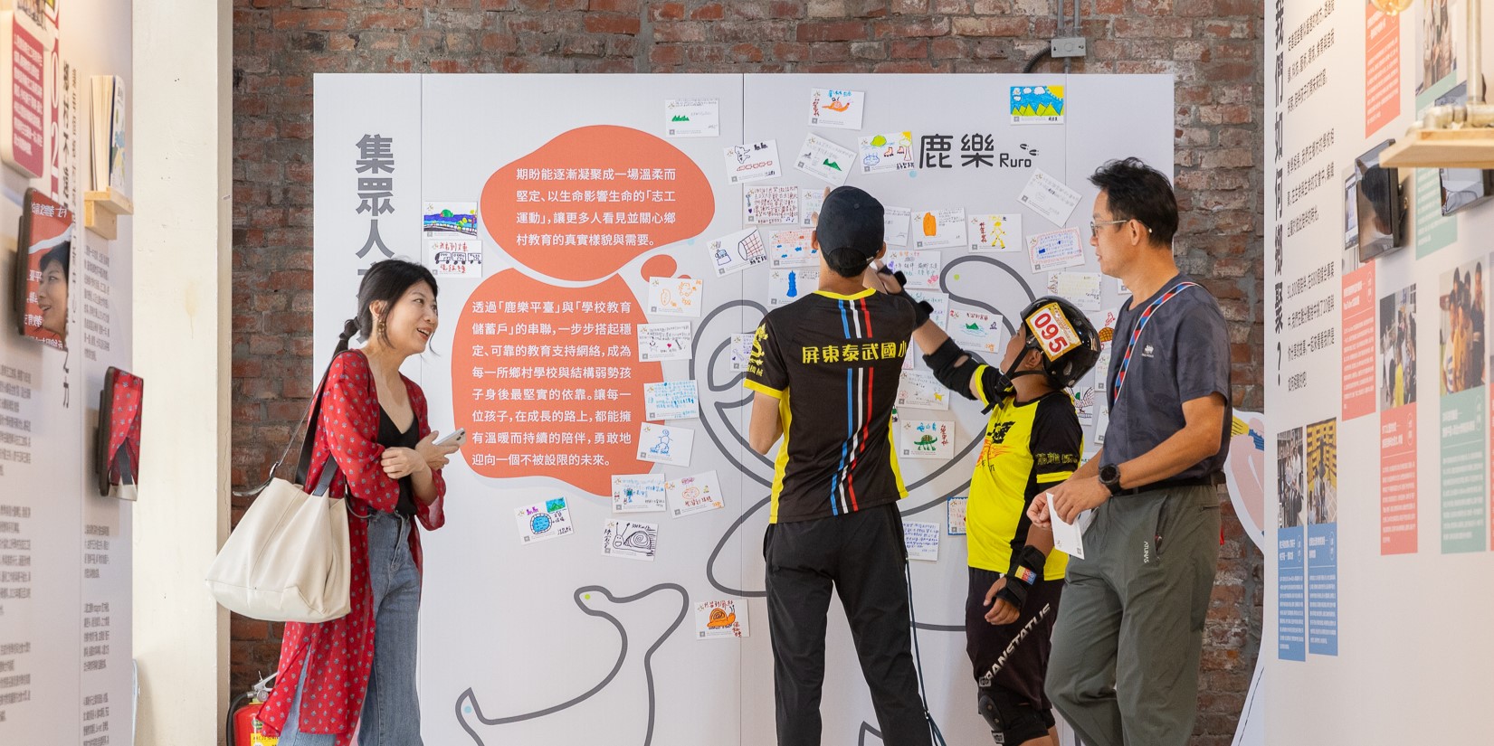 展覽現場，一名女性左側觀看海報，右側三位騎行者穿著黑黃色車衣停留在「鹿樂Ruro」主題展板前。展板貼滿手繪圖案與文字說明，中央橘色對話框介紹偏鄉教育理念，背景為紅磚牆。