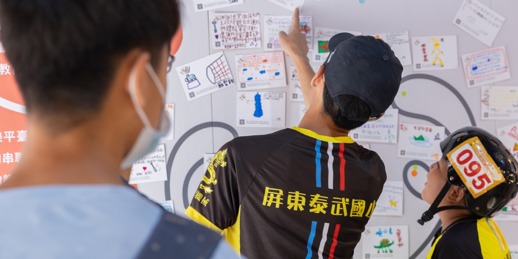 兩名穿著黑黃色運動服的人正在指向貼滿白牆的各式設計草圖和創意方案。牆上展示著多個手繪圖表、色彩配置和產品設計稿，右側人物戴著安全帽。這是一場設計評審或創意工作會議的場景。