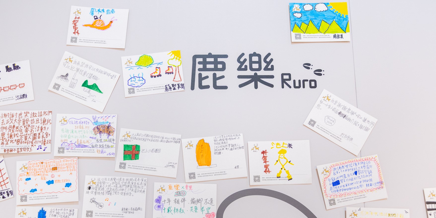 中央顯示「麗樂 Ruro」文字，周圍散放多張兒童明信片。明信片上有手繪圖案、彩色插畫和日文書寫內容，包括太陽、山景、房屋、人物等主題。每張卡片右上角有QR碼，展示兒童美術創作和郵寄活動成果。
