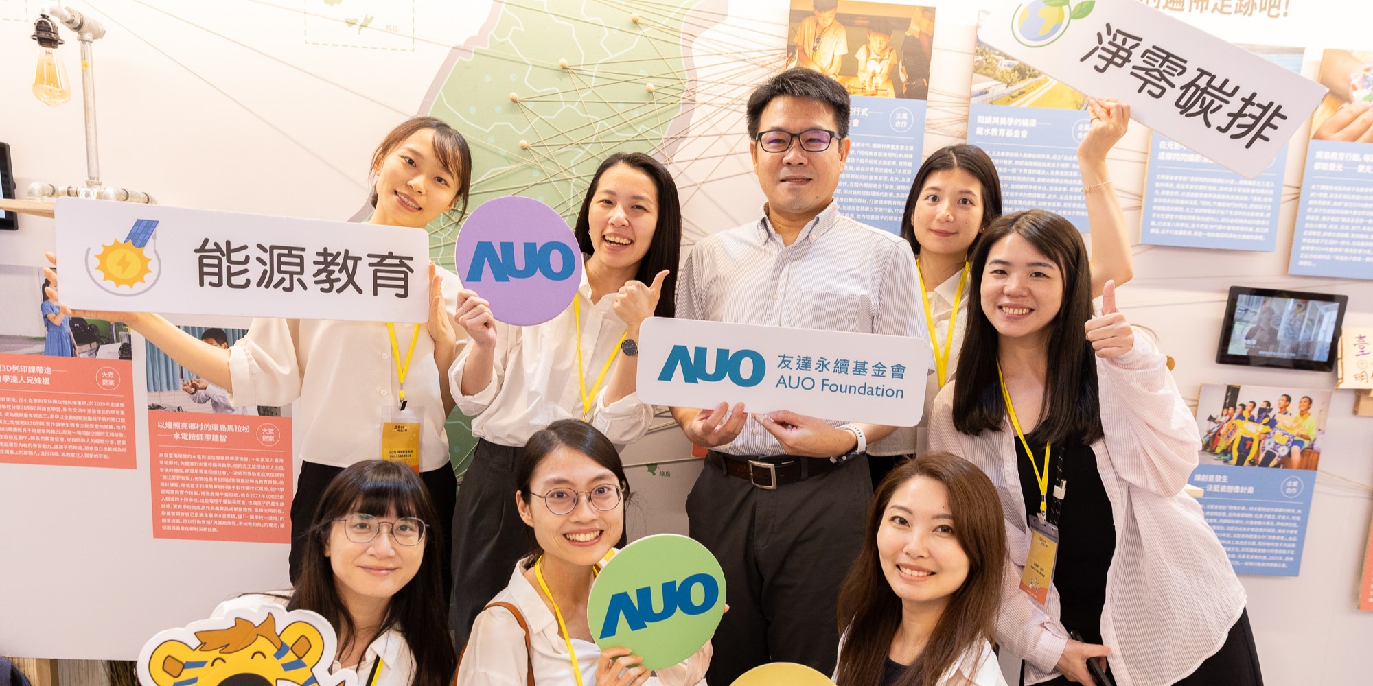 九位佩戴識別證的年輕人在展覽會場合影，展示能源教育、AUO及友達永續基金會的標誌。背景為教育推廣展示板，牆上張貼中文海報及相關資訊。團隊成員穿著白色襯衫，表情親切歡樂，呈現教育推廣活動的現場氛圍。