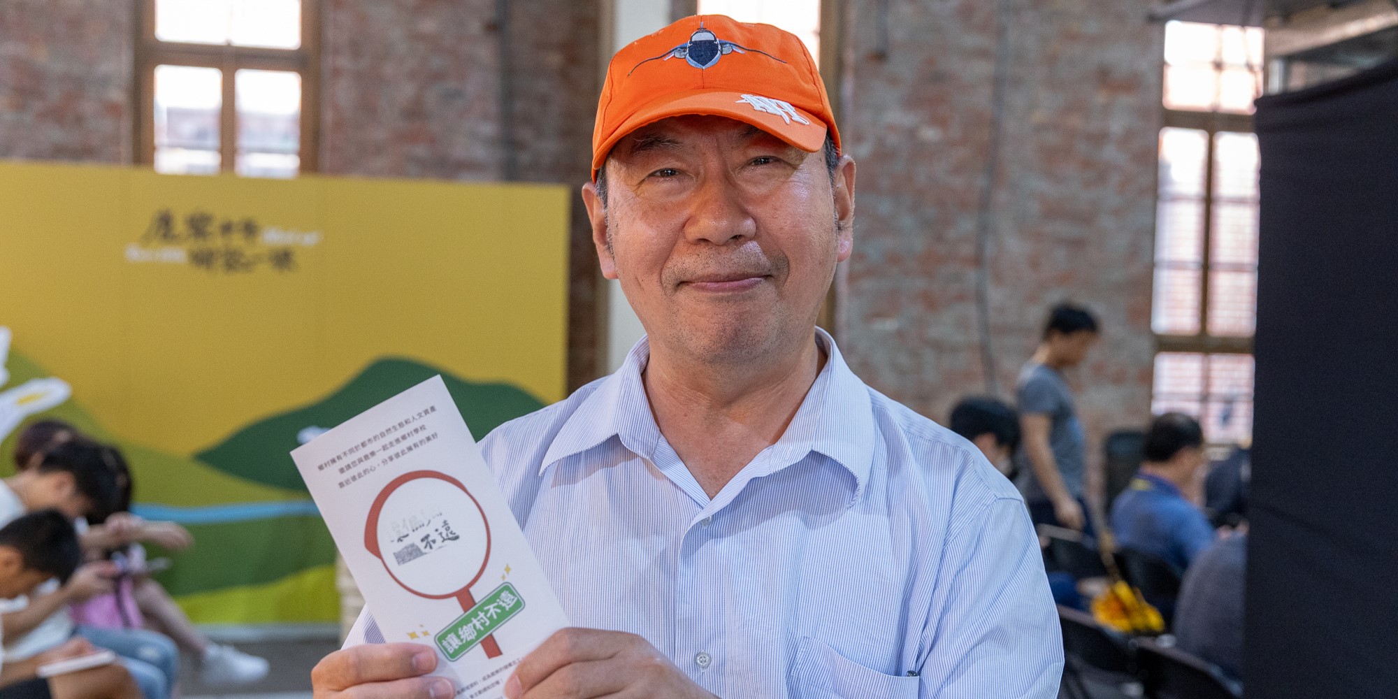 一位中年男性穿著淺藍色襯衫和橙色棒球帽，在室內活動現場展示一份宣傳單張。背景有黃綠色海報和磚牆，多名參與者在場。男性面帶微笑，展示的文件上有紅色圓形標記和中文字樣。