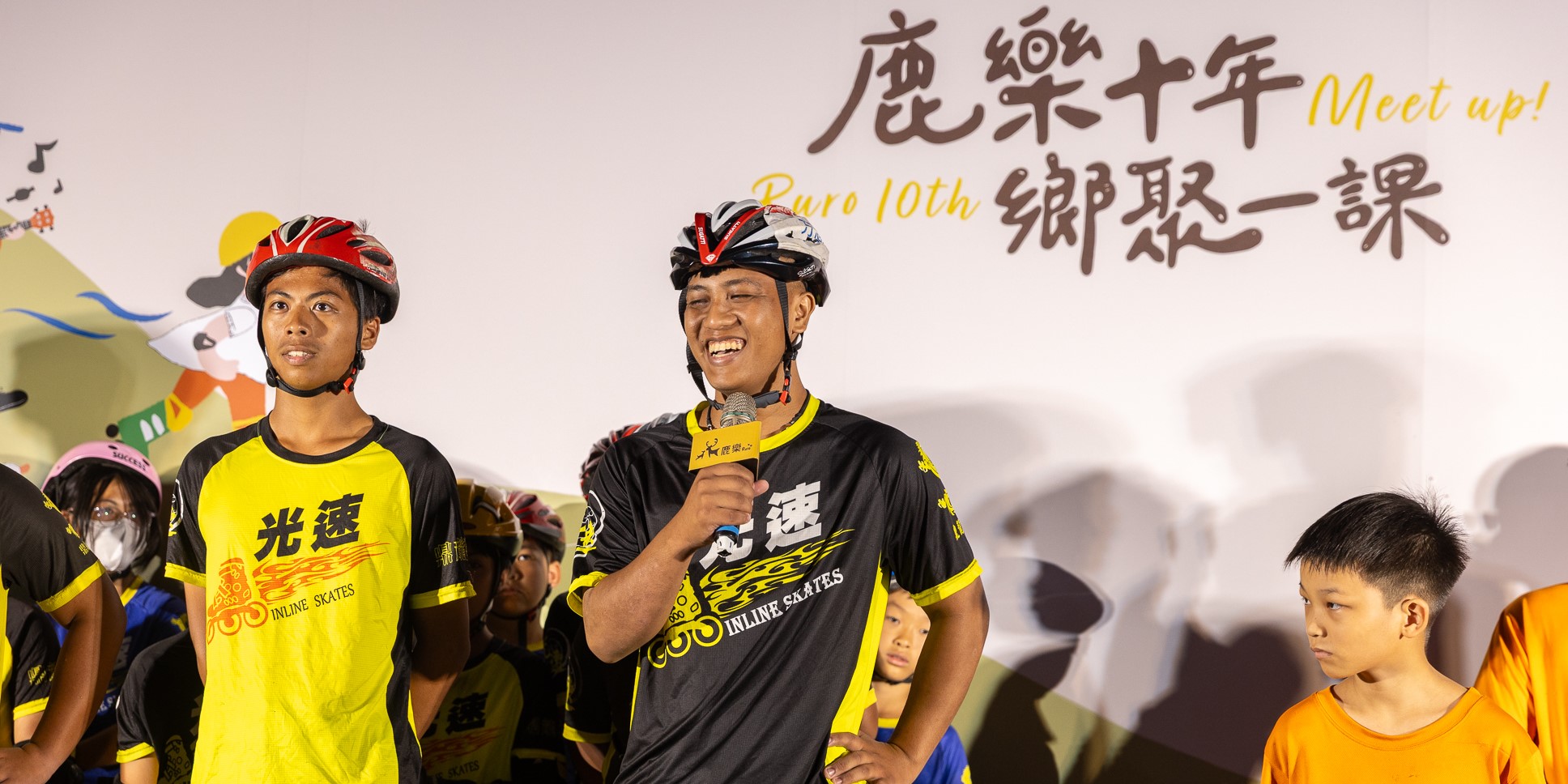兩名穿著黃黑色輪滑隊服、戴著安全帽的年輕男性運動員在舞台上，其中一人拿著麥克風發言。背景牆上寫著「麗泉十年」、「Meet up!」、「Buro 10th」和「鄰聚一課」的中英文文字。舞台下有兒童觀眾。