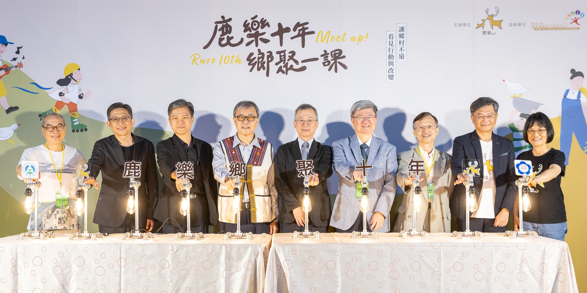 鹿樂十年鄉聚一課活動現場，十位與會者手持點亮的燈籠站成一排，背景為活動主視覺與卡通人物插圖，象徵點亮鄉村教育的承諾與團結。