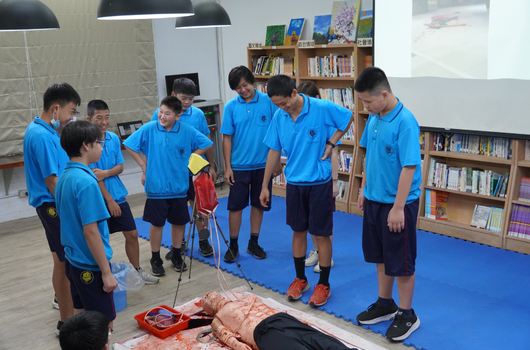 一群穿著藍色制服的學生在圖書館內進行急救訓練。一名學生躺在地上模擬患者，其他學生圍成一圈觀看和學習心肺復甦術的正確操作步驟。教室背景可見書架和投影螢幕。