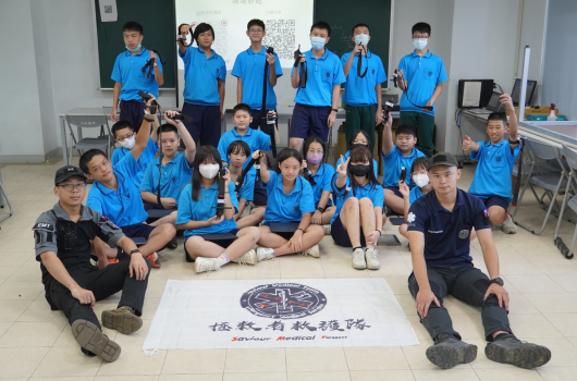 一群穿著藍色運動服的學生在教室內合影，圍繞著寫有校隊標誌的白色橫幅。兩位教練穿著黑色運動服坐在前方兩側。學生們有站有坐，呈現團隊凝聚力的合照場景。