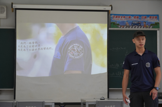 教室內，一位穿著深藍色制服的年輕男性站在投影幕前進行簡報。投影幕上顯示警察制服袖章和中文文字說明。背景黑板上可見教學內容。這是一場關於警察制度或執法工作的教育演講。