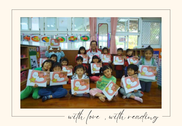 幼兒園教室內，一位女性教師與約二十位幼童坐在木質地板上，每位幼童手持自己繪製的彩色圖畫，開心地面向鏡頭。圖片下方標語為「with love, with reading」。