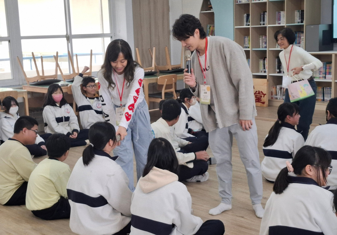 學校圖書館內，兩位配戴識別證的成人講師向坐在地上的約二十位學生進行教學互動。學生穿著白黑相間的制服，講師之一手持麥克風進行講解。背景為書架、木製椅子和大型窗戶。