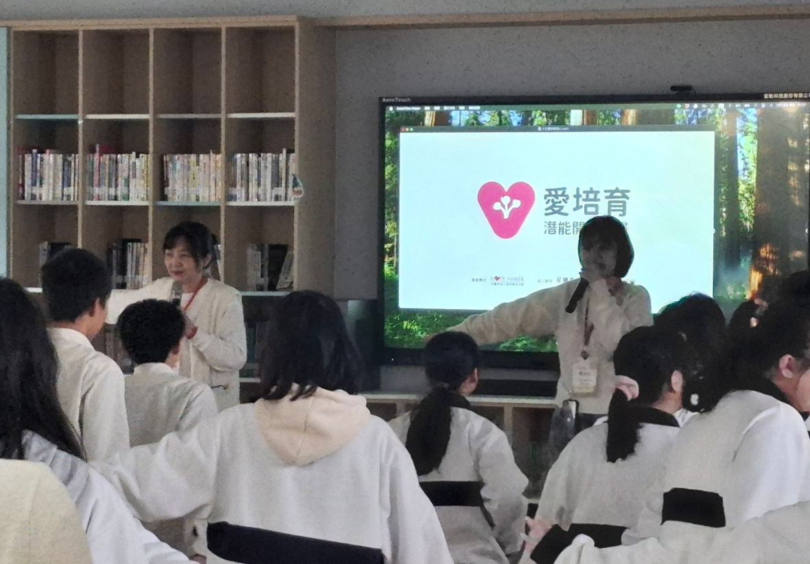 教室內舉辦講座活動，約十多名穿著校服的學生坐在座位上聽講。講台上有兩位講者，大螢幕顯示粉紅色愛心標誌和「愛培育」文字的簡報。背景為木製書架，擺放多排書籍。
