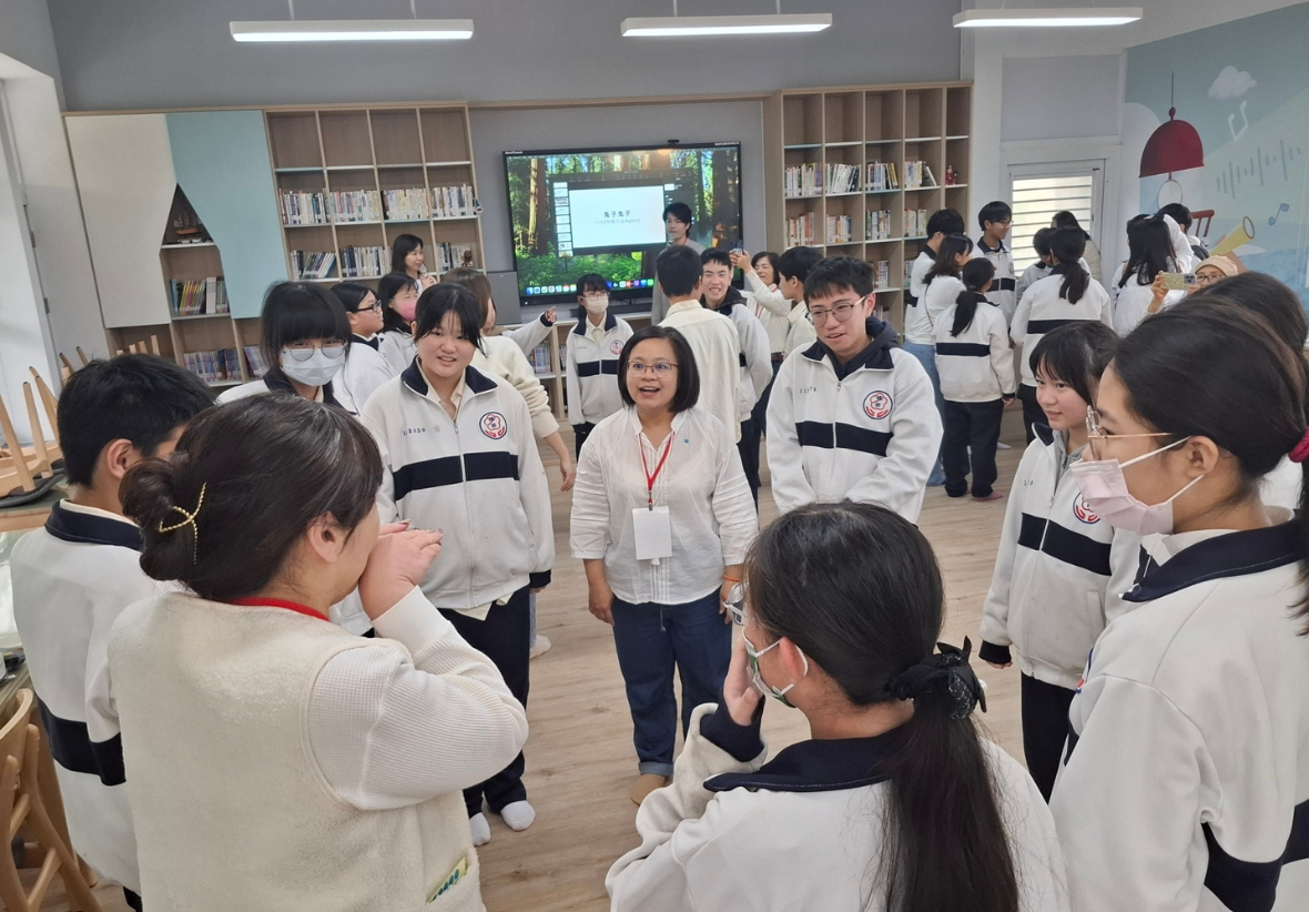 一群穿著白色黑條紋校服的學生在圖書館內聚集，中央一位配戴識別證的女性講者正在進行校園活動或教學。背景展示書架、電視螢幕及溫暖的木質裝潢。學生們都戴著口罩，認真聆聽講解。