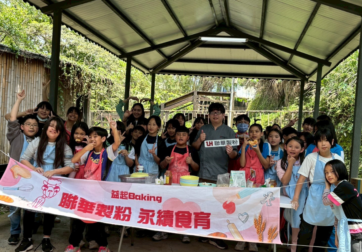 一群學生在戶外棚架下進行烘焙活動，舞台前方展示寫有「聯華製粉 益起Baking 永續食育」的粉紅色橫幅，參與者穿著藍色圍裙，舉手歡呼，現場擺放烘焙工具與材料，展現食育教育推廣活動的熱鬧氛圍。