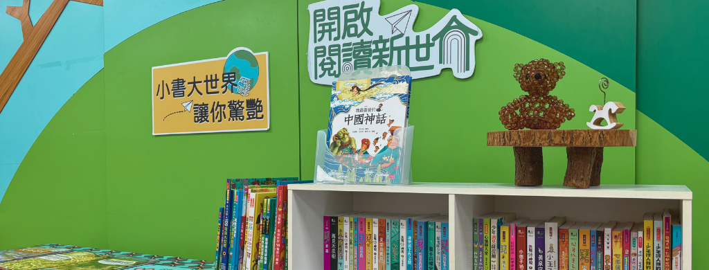 兒童閱讀區展示牆，綠色背景上有「小書大世界讓你驚豔」和「開啟閱讀新世代」標語，牆面裝飾有樹木和搖馬圖案。下方白色書架整齊排列著數百本彩色兒童書籍，左側地面散放幾本打開的圖書。
