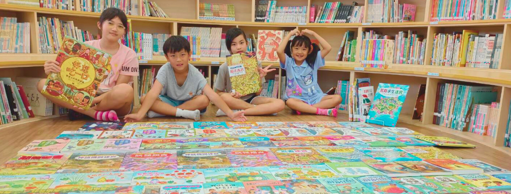 四名兒童坐在圖書館木製地板上，身後是書櫃。他們展示著五本色彩繽紛的兒童繪本，書籍鋪散在地面上。孩子們穿著粉紅、藍色等淺色衣服，開心地擺姿勢拍照，營造溫馨的閱讀推廣氛圍。