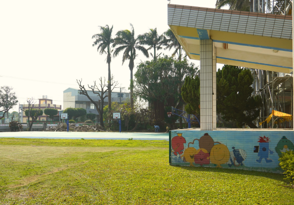 公園草坪上，右側黃藍色建築物牆面繪有卡通人物和房屋圖案。背景可見棕櫚樹、工業建築和晴朗天空。寬敞綠色草地供遊憩使用。