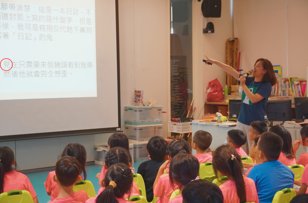 教室內，一位女老師站在講台上，用麥克風向坐在黃色椅子上的小學生講授中文課程。投影幕上顯示含有繁體中文的教學內容。教室後方擺放書籍和教材。學生認真聆聽老師的講解。