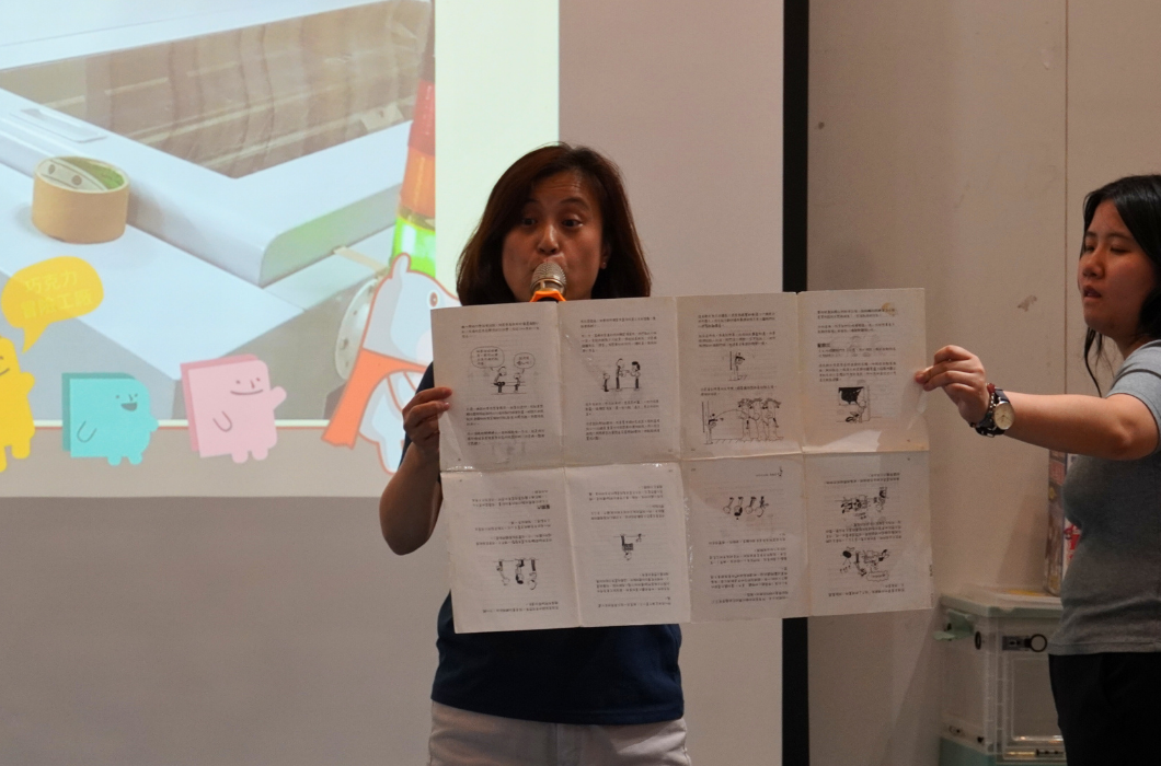 一位女性在演講時展示一份多頁的說明書或教材，頁面上有插圖和文字說明。身旁另一位女性協助展示內容。背後投影幕上顯示卡通角色和物品的插圖，呈現教育或產品介紹的演講場景。