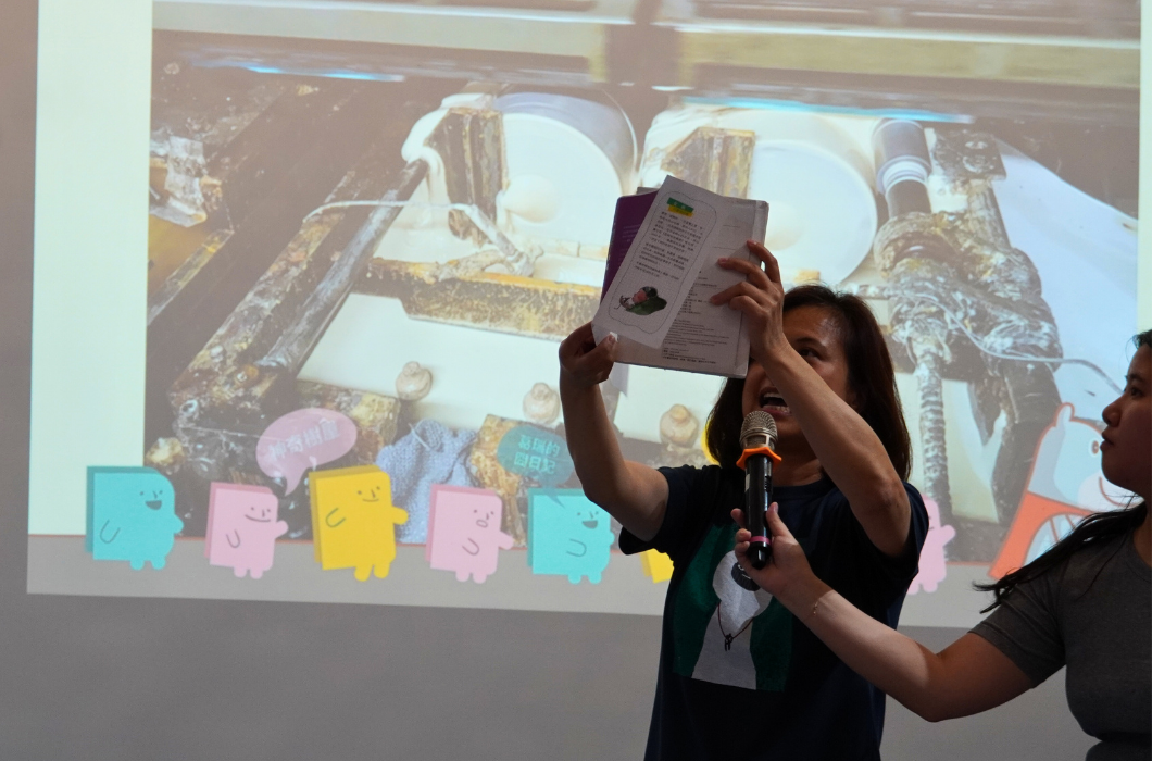一位女性在舞台上手舉書籍，持著麥克風進行演講。身後投影幕顯示太空船艙內景象及彩色卡通角色。舞台下方有工作人員協助。這是一場兒童教育或科普主題的活動演講現場。