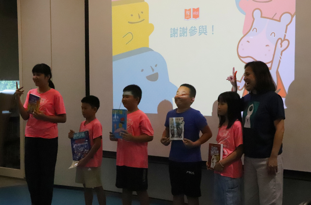一位女性主持人與五位兒童站在投影螢幕前，兒童穿著紅色或藍色T恤，手持書籍或獎狀。背景投影顯示卡通人物和中文文字「謝謝參與」，呈現兒童閱讀表揚活動的場景。
