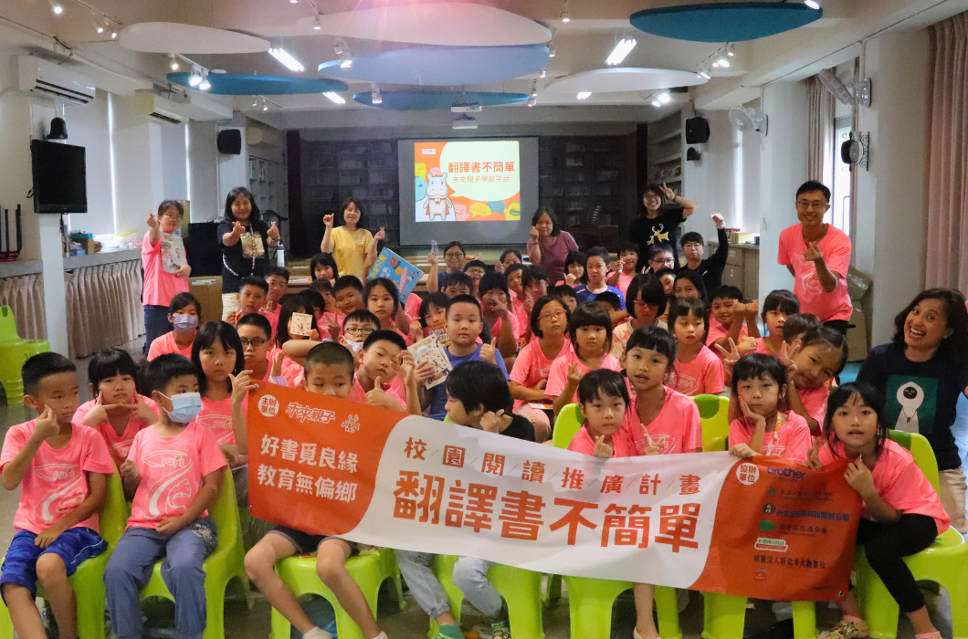 一群穿著粉紅色T恤的兒童在室內聚集，舉著寫有「翻譯書不簡單」和「好書sight閱線教育無偏鄉」的宣傳標語。舞台上播放著相關推廣影片，多位成人志工在場協助活動進行。這是一場推廣閱讀教育的校園活動。