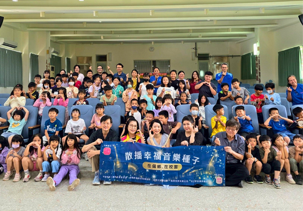 大型室內活動現場，約百位大人和小孩齊聚一堂，前排舉著藍色布幡寫著「散播幸福音樂種子」，參與者穿著整齊坐成多排，展現溫馨的社區或教育機構集體活動情景。
