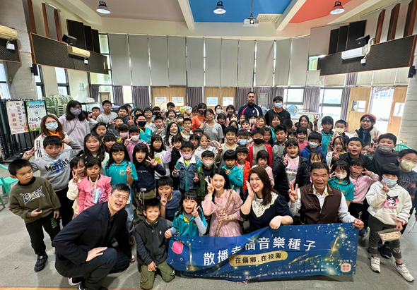 小學師生與來賓在校園室內合照，眾人聚集在一起舉著寫有「散播音樂種子」的藍色布幡，慶祝音樂教育推廣活動。天花板為藍色和紅色設計，窗戶採光良好，參與者包括兒童和成人，多數配戴口罩。